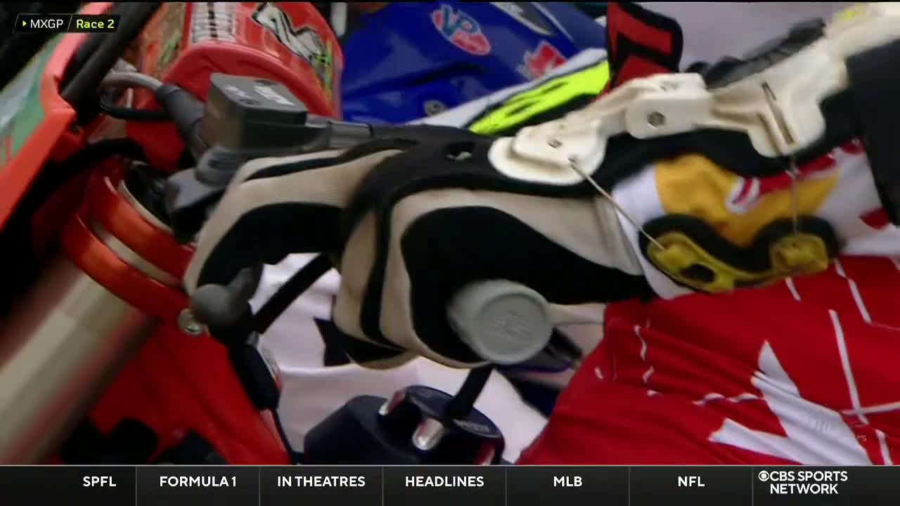 A gloved hand grips the handlebar, ready to twist the throttle. The rider's gear, red and white, is visible as the CBS Sports Network USA broadcast focuses on the start of MXGP Race 2.
