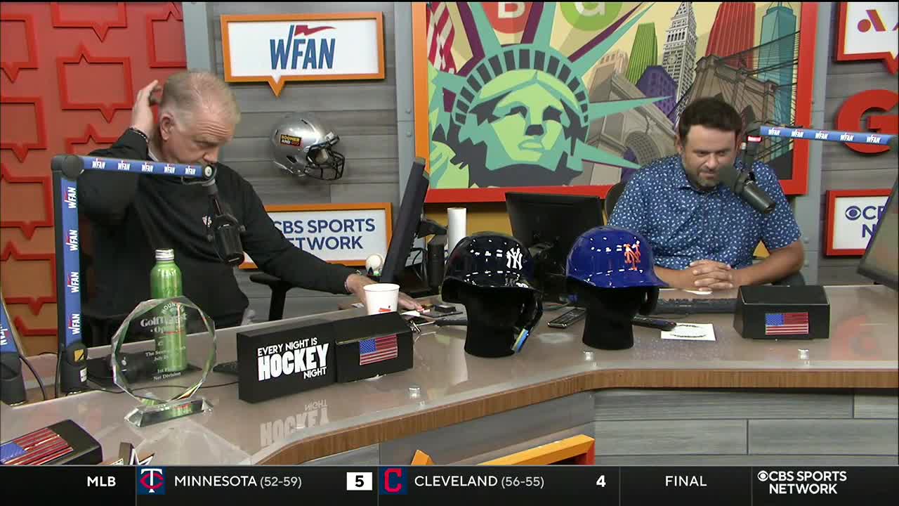 Two men sit behind a desk, discussing something on CBS Sports Network USA. Baseball helmets sit in a row, and the man on the left is adjusting his hair.
