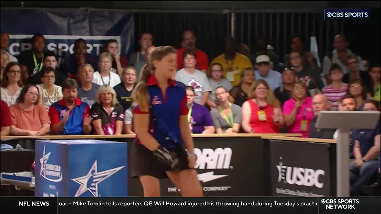 A bowler in a red and blue shirt stands at the lane, holding her bowling ball. The crowd behind her, watching the action on CBS Sports Network USA, is applauding.
