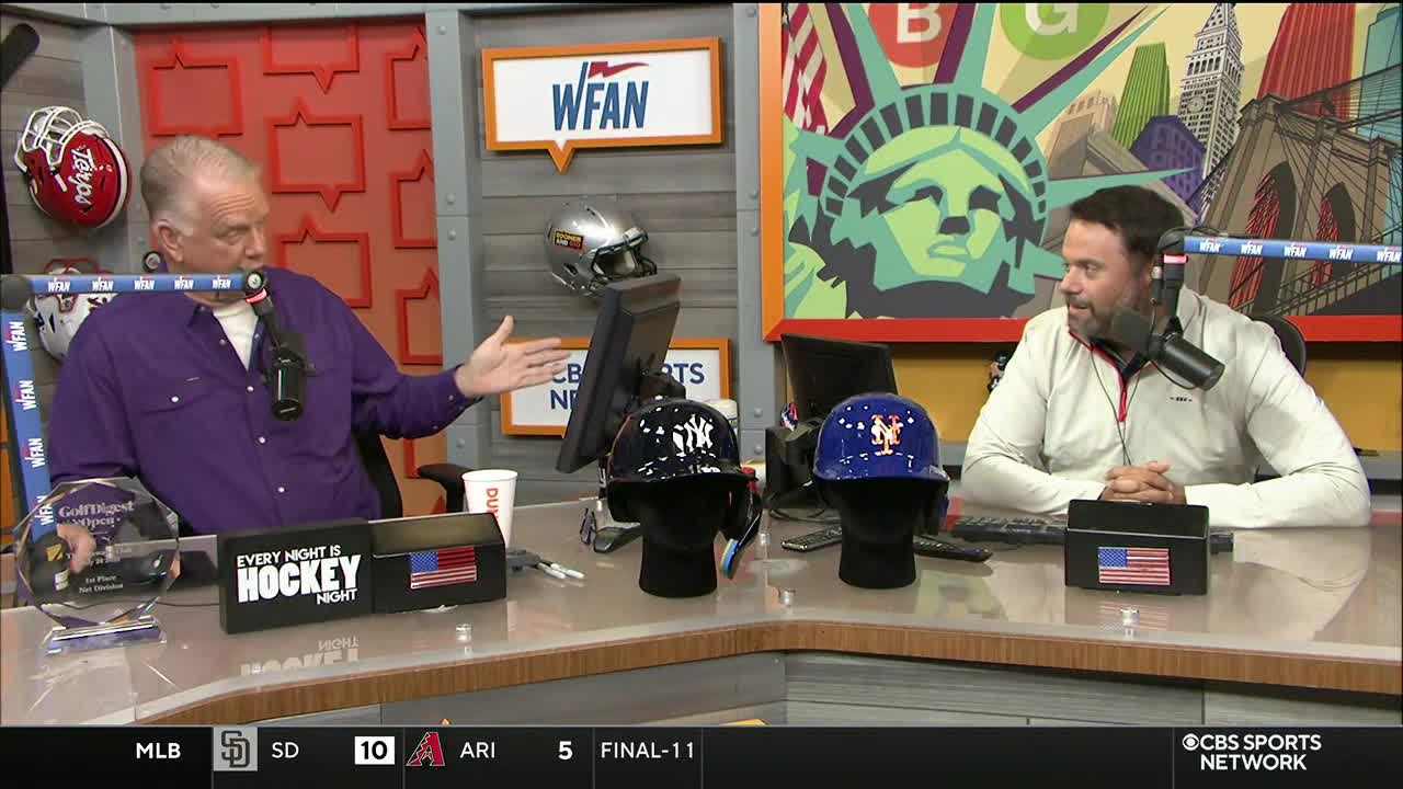 Two men are seated at a desk, discussing sports on CBS Sports Network USA, with baseball helmets on display between them. The man in the purple shirt gestures towards the helmets while the other man listens with a microphone.
