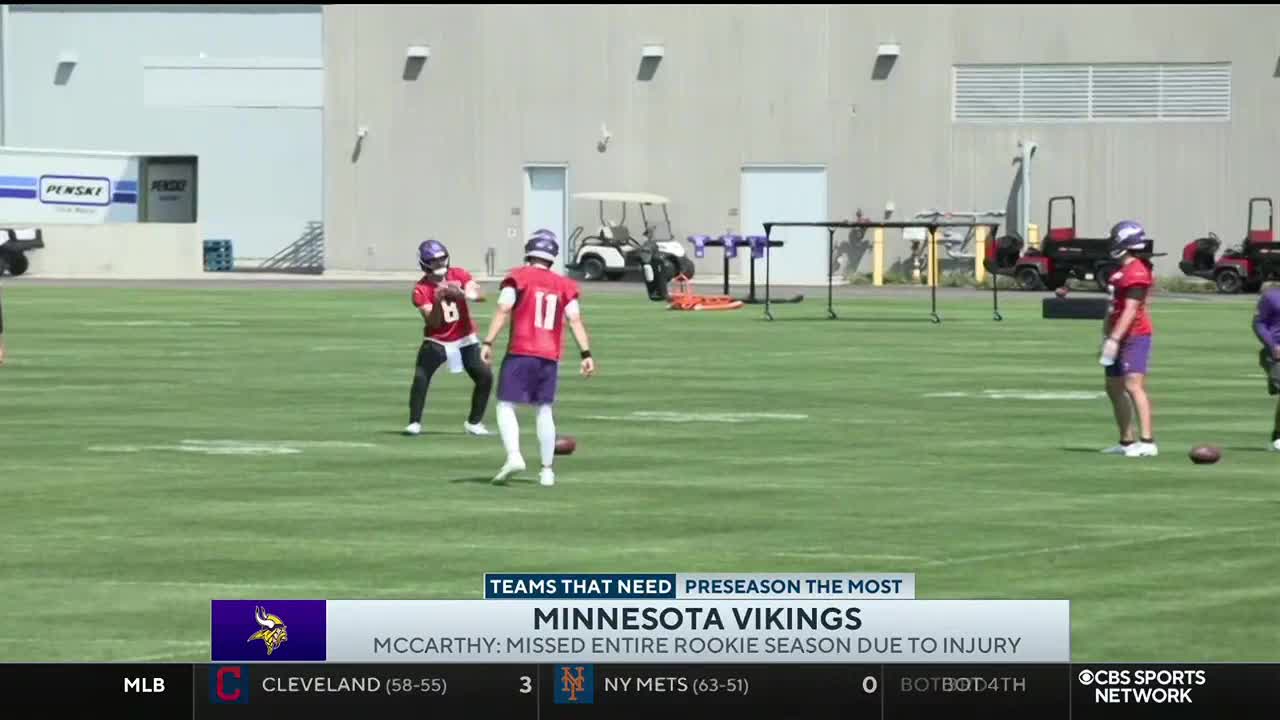 On a sunlit field, Minnesota Vikings players in purple and red practice. A quarterback, number 11, is about to throw the ball.
