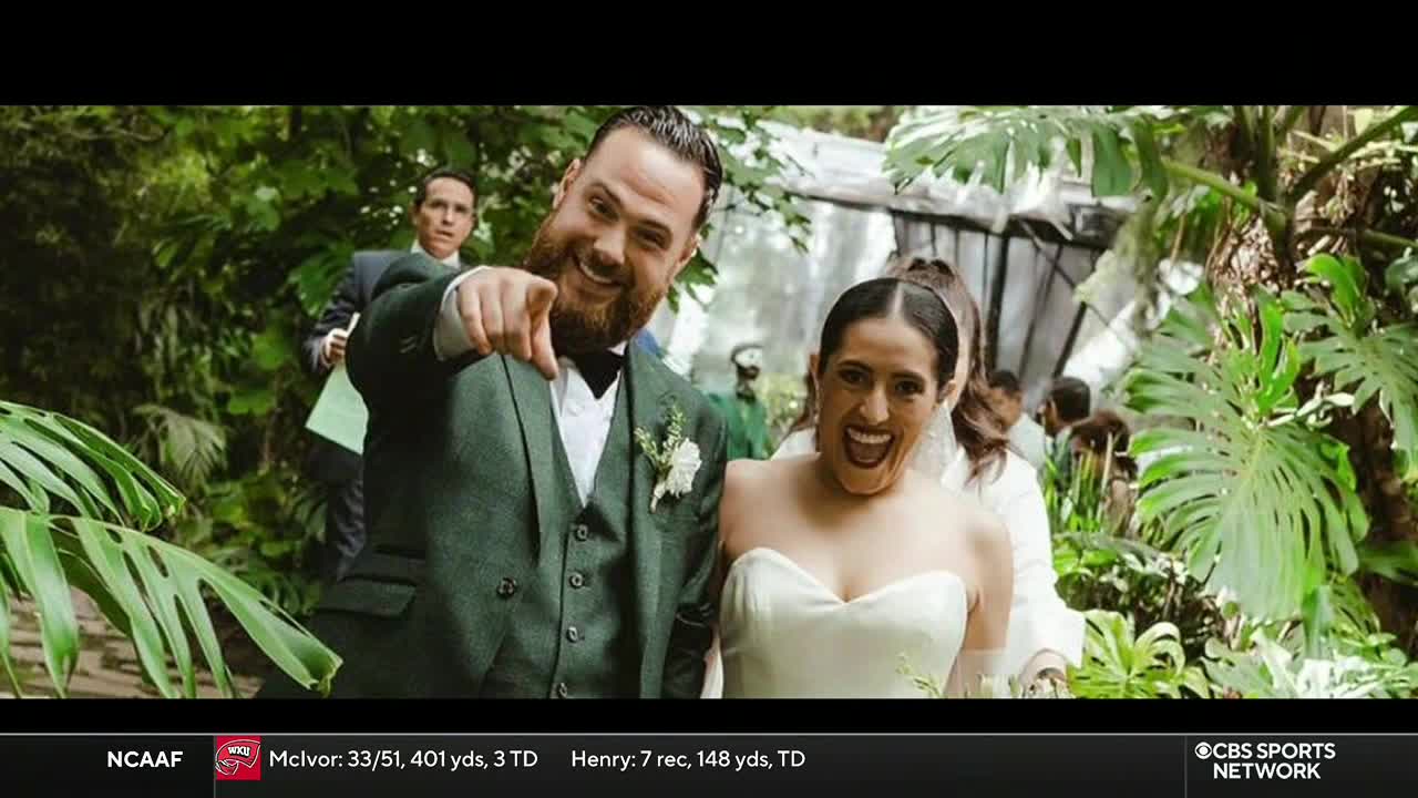 A beaming groom, in a green suit, points directly at the camera, while his bride grins widely beside him. Lush greenery surrounds them, and a man in a suit stands behind them, holding a paper.
