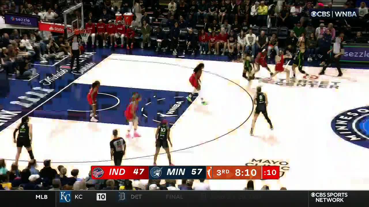 The Indiana Fever in red are on offense, with the ball being dribbled up the court. The Minnesota Lynx, in black and green, are trying to defend near the free throw line.
