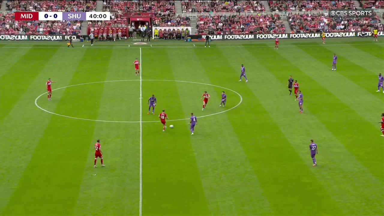 The player in red dribbles the ball toward the center circle, pursued by a player in purple. A few other players from both teams are positioned further upfield, ready to receive a pass.
