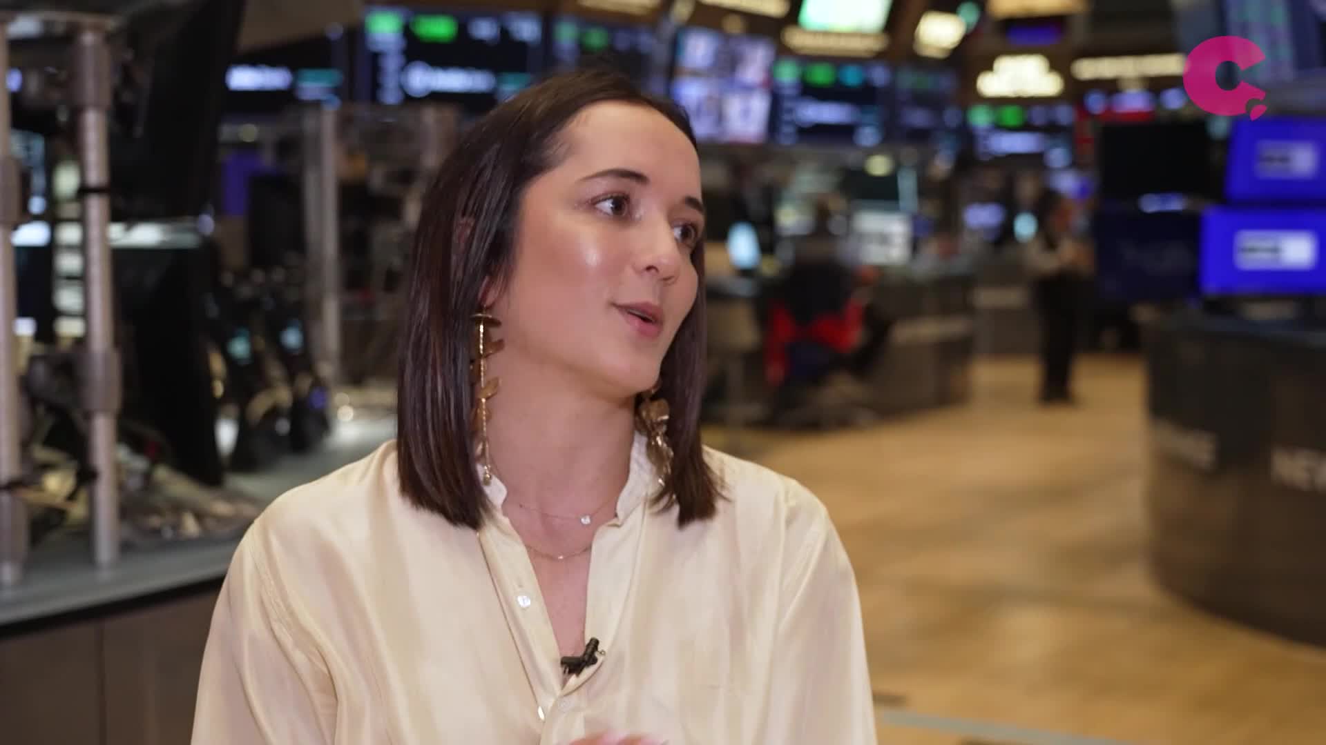 A woman in a cream-colored blouse speaks, her gaze directed slightly to the side. Behind her, the screens and bustle of a trading floor are visible, with the Cheddar News logo in the corner.

