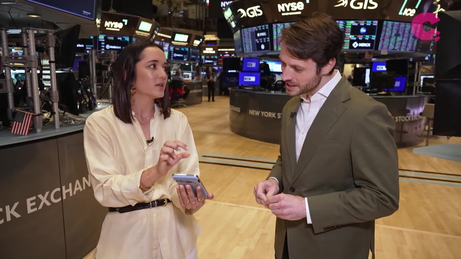 A woman in a cream-colored shirt gestures with her right hand while holding a phone, speaking to a man in a green blazer. They're standing in the New York Stock Exchange, likely reporting for Cheddar News.
