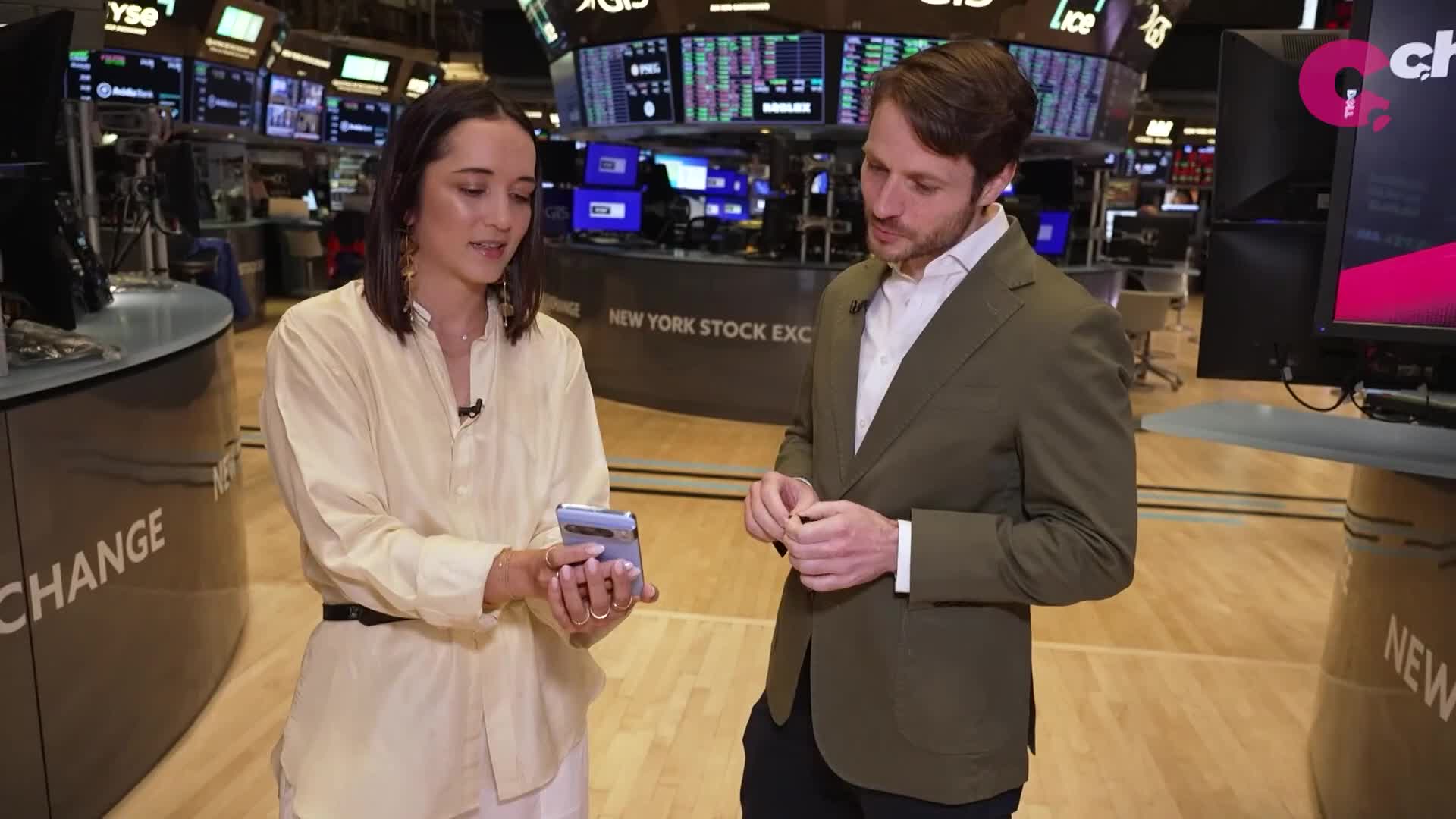 A woman in a light yellow shirt holds a phone, gesturing as she speaks to a man in a green blazer. The pair stands in front of the New York Stock Exchange, with screens displaying data in the background.
