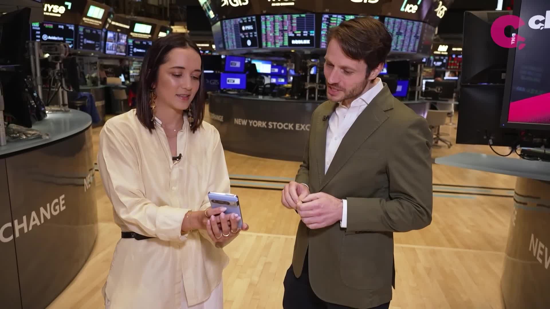 A woman in a light yellow shirt holds a phone, pointing at the screen while speaking to a man in a green blazer. They're standing in front of screens displaying stock information, likely on a set for Cheddar News in the United States.
