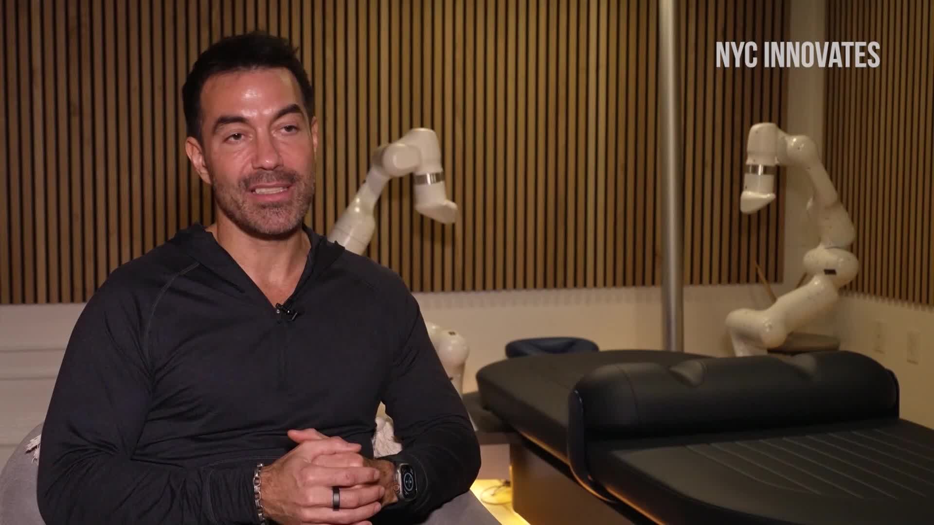 A man in a black shirt is talking, his hands clasped in his lap. Behind him, two white robotic arms hover over what looks like massage tables.
