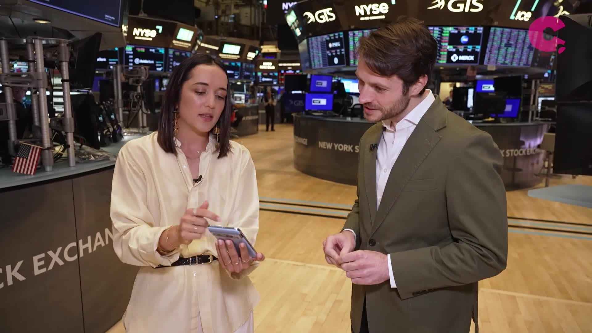 A woman in a cream-colored shirt gestures at a phone, speaking to a man in a green blazer. They're standing on the floor of the New York Stock Exchange, with screens displaying market data in the background.
