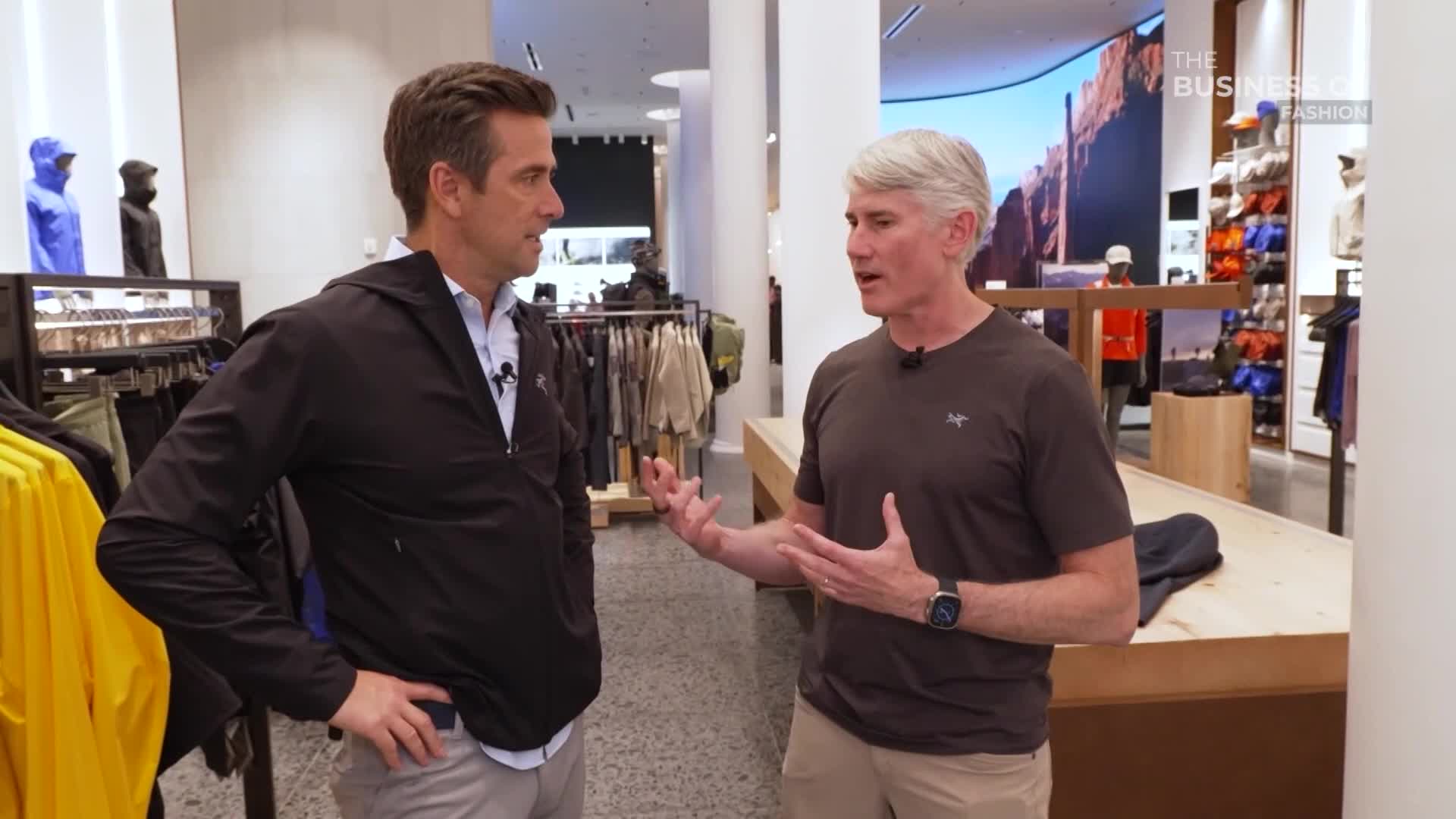 Two men are in a retail store, one gesturing animatedly with both hands while speaking. The other man, wearing a black jacket, listens with his hand on his hip.
