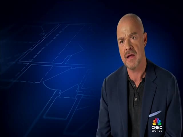 A man in a blazer, with a CNBC World logo, is speaking directly to the camera against a blue background with a blueprint design. He's gesturing with his hands as he talks.
