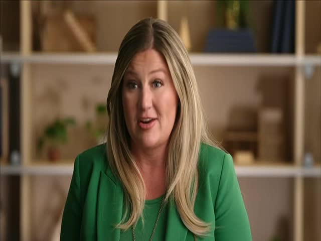 A woman in a green blazer is speaking directly to the camera, her expression open and engaged. Behind her, a simple wooden shelving unit provides a neutral backdrop.
