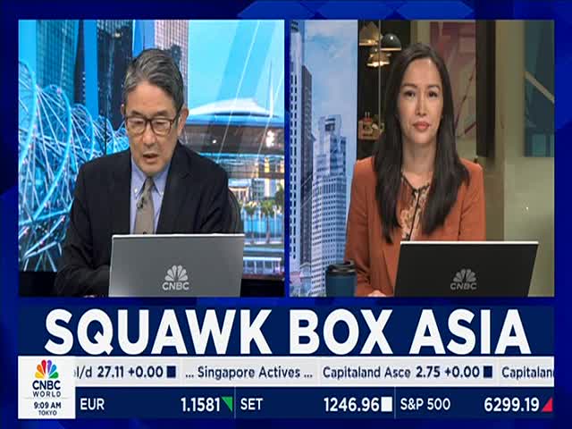 Two anchors sit at their desks, working on laptops, with a backdrop of cityscapes. The "Squawk Box Asia" logo and market data are displayed on the screen below them, broadcast by CNBC World.
