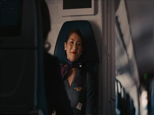 A smiling flight attendant in a dark uniform sits in an airplane seat, her red lipstick standing out. Across the aisle, the back of another passenger is visible.
