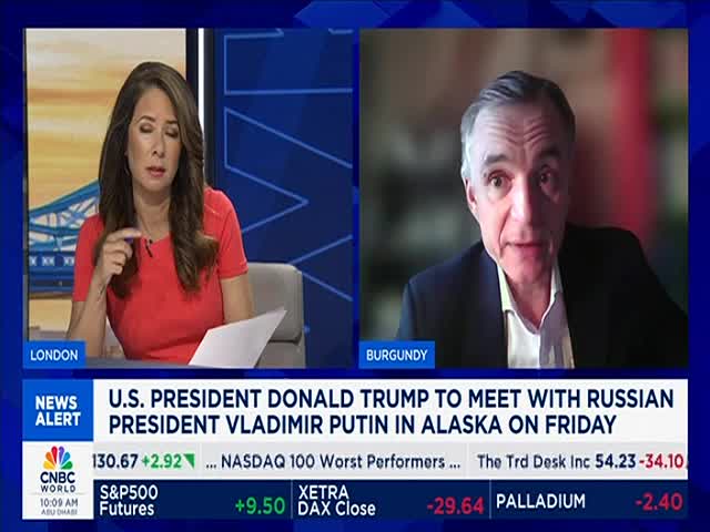 A presenter in a red top gestures while looking at a document. Across the screen, a man in a dark suit speaks from Burgundy, likely on CNBC World, as a news alert flashes.
