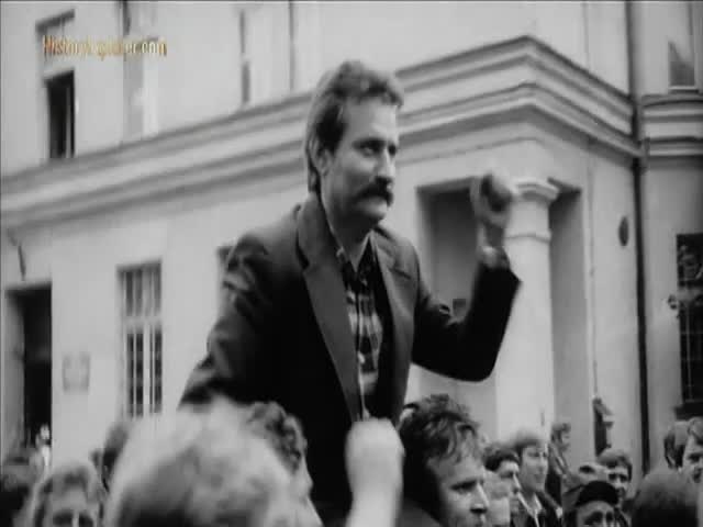 A man with a thick mustache is raised above the crowd, his fist clenched in the air. He's wearing a jacket and a plaid shirt, and the people below are looking up at him.
