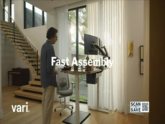 A man stands next to a standing desk, looking down at it. The desk has two monitors mounted on it, and the words "Fast Assembly" are superimposed over the scene.
