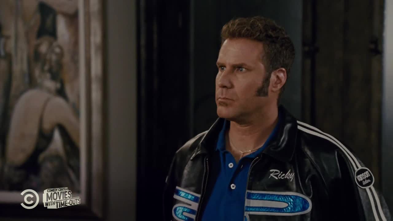 The man in the racing jacket is looking off to the side, his face set in a serious expression. The Comedy Central logo is in the corner, and a painting is visible behind him.
