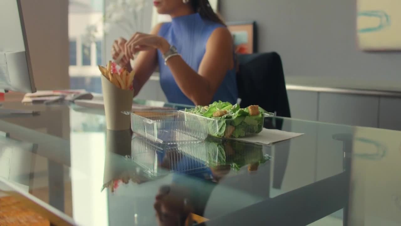 A woman in a blue top sits at a glass desk, with a container of salad and a cup of fries nearby. She's in an office, likely in the United States, and appears to be working or taking a break.
