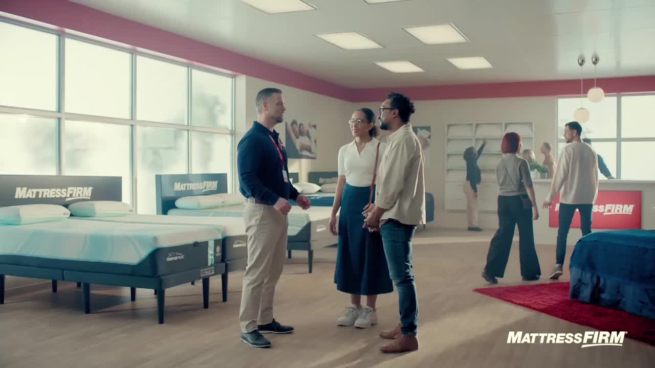 A salesperson in a dark jacket gestures toward a couple, who stand facing him in a Mattress Firm store. Other customers wander around, looking at the beds.
