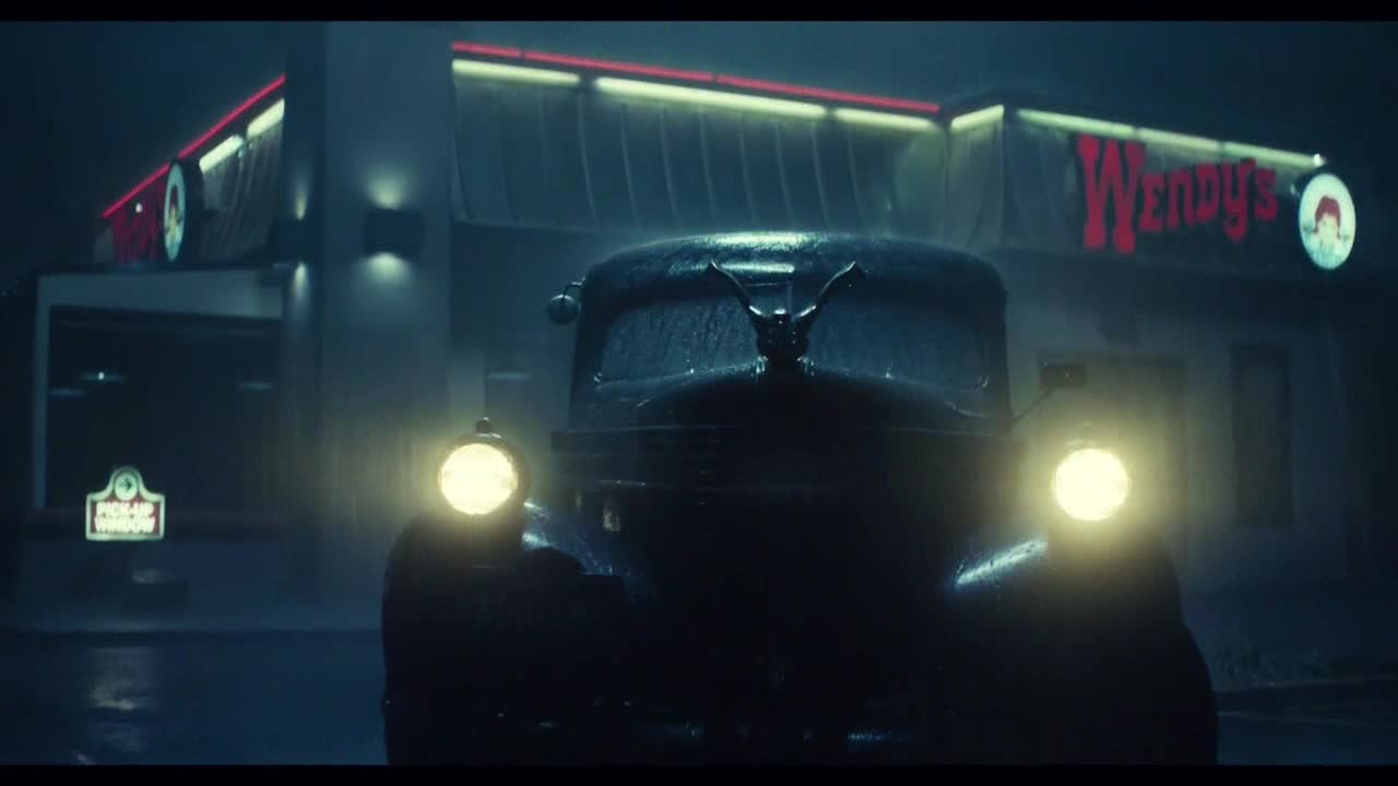 A classic car, its headlights glowing, sits in the rain in front of a Wendy's. The red neon sign and the fast food restaurant's logo are visible in the background.

