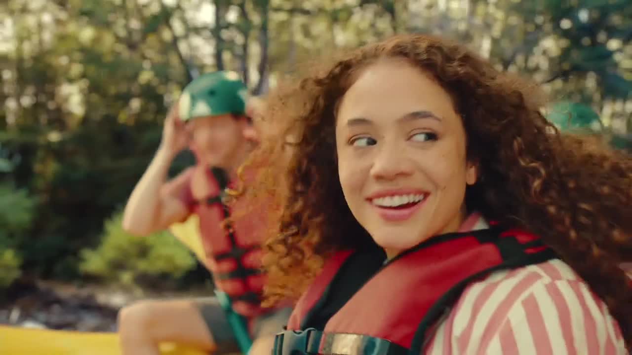 A woman with curly hair grins, looking over her shoulder as she rides in a boat. Behind her, a person in a green helmet adjusts it, also wearing a life vest.
