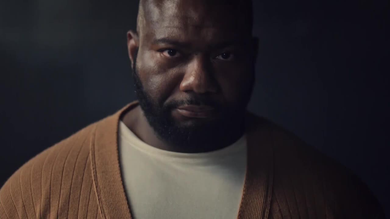 A man with a shaved head and a full beard stares directly ahead, his expression intense. He's wearing a light-colored shirt and a tan cardigan, the lighting casting shadows on his face.

