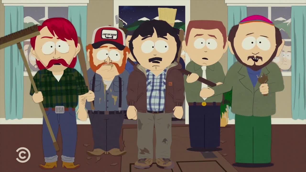 A group of men, some holding brooms and sticks, surround a man with a brown jacket. He stands with a bewildered expression, as if he's not sure what's going on.
