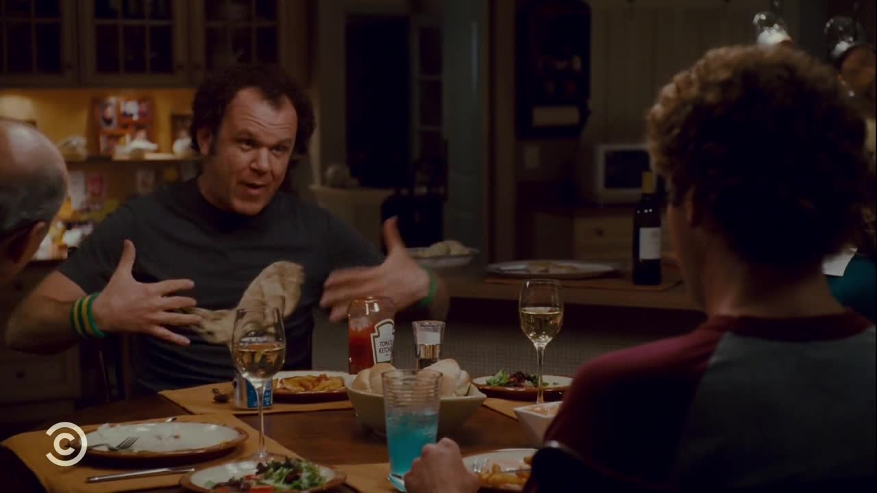A man in a gray t-shirt gestures emphatically, his hands splayed wide as he speaks at the dinner table. Beside him, someone with curly hair listens intently, the red and gray of their shirt visible.

