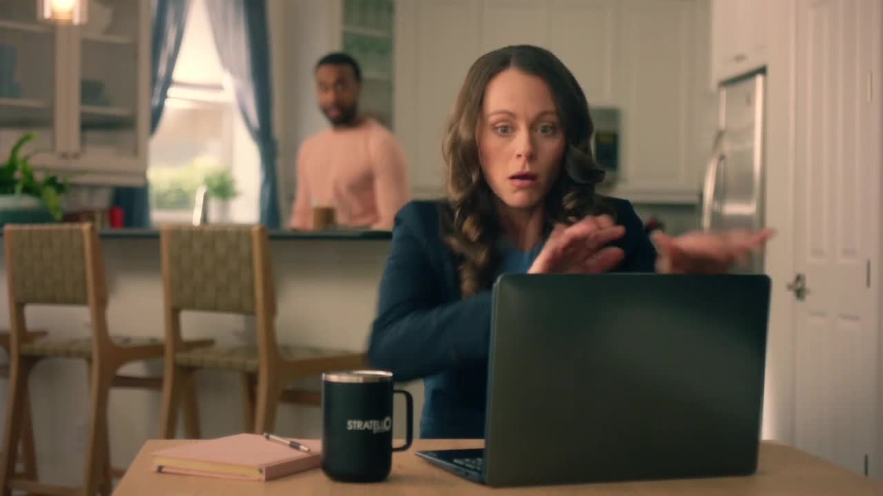 The woman's eyes are wide, and her hands are raised in front of the laptop, as if she's reacting to something onscreen. A man stands in the background, looking on.
