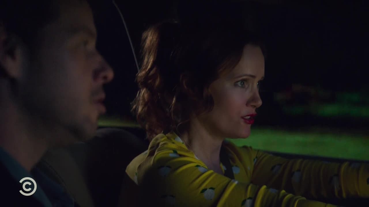 Inside a car at night, a woman with red lipstick and a yellow patterned shirt is talking animatedly. The man next to her listens intently, his face illuminated by the car's interior light.
