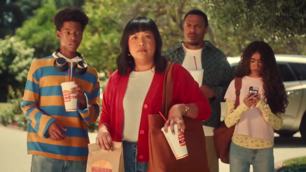 A family, carrying Burger King cups and bags, walks toward the camera. The woman in the red cardigan looks concerned while her daughter is engrossed in her phone.

