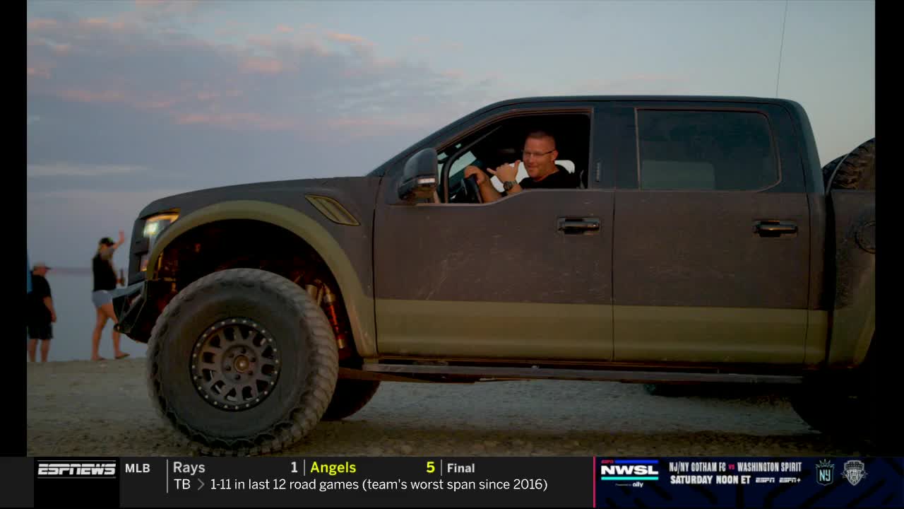 A man in a dark truck, wearing glasses, waves from the driver's seat. Two people stand near the water in the background, while the ESPNews logo is visible in the lower left corner.
