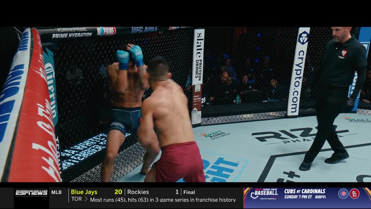 A fighter in blue gloves and shorts is backed against the cage, hands up. The other, in red shorts, is winding up for a punch as the referee watches.
