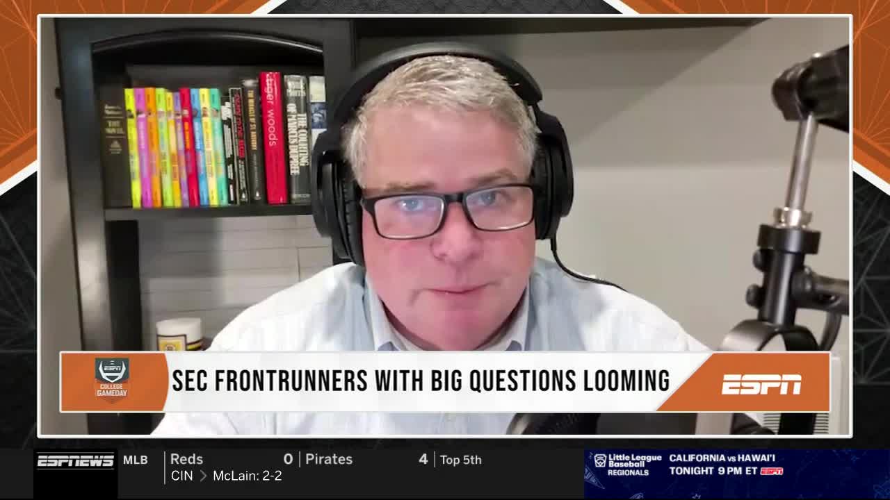 A man with glasses and headphones is speaking directly into the camera, framed by the ESPN logo and a graphic about SEC frontrunners. Behind him, a bookshelf holds a collection of books and colorful highlighters.
