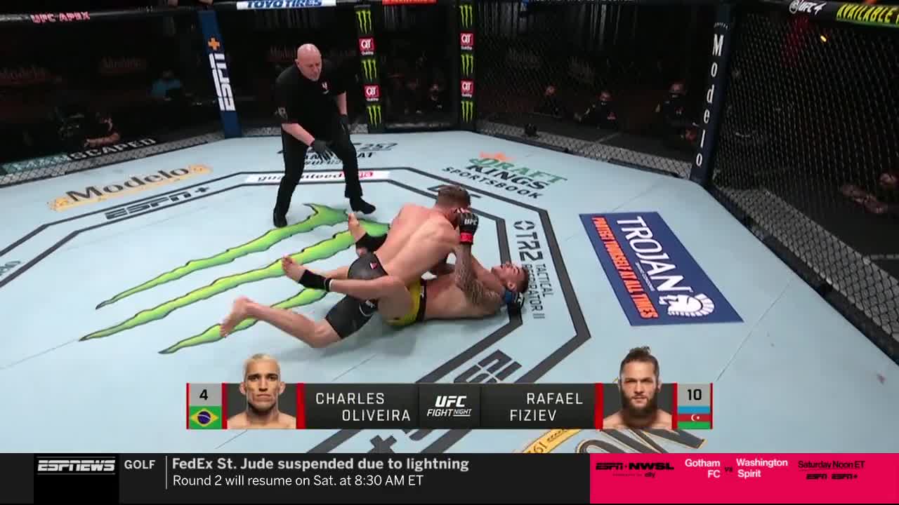 Charles Oliveira has Rafael Fiziev pinned on the mat, grappling. The referee stands nearby, watching the action unfold on ESPNews.
