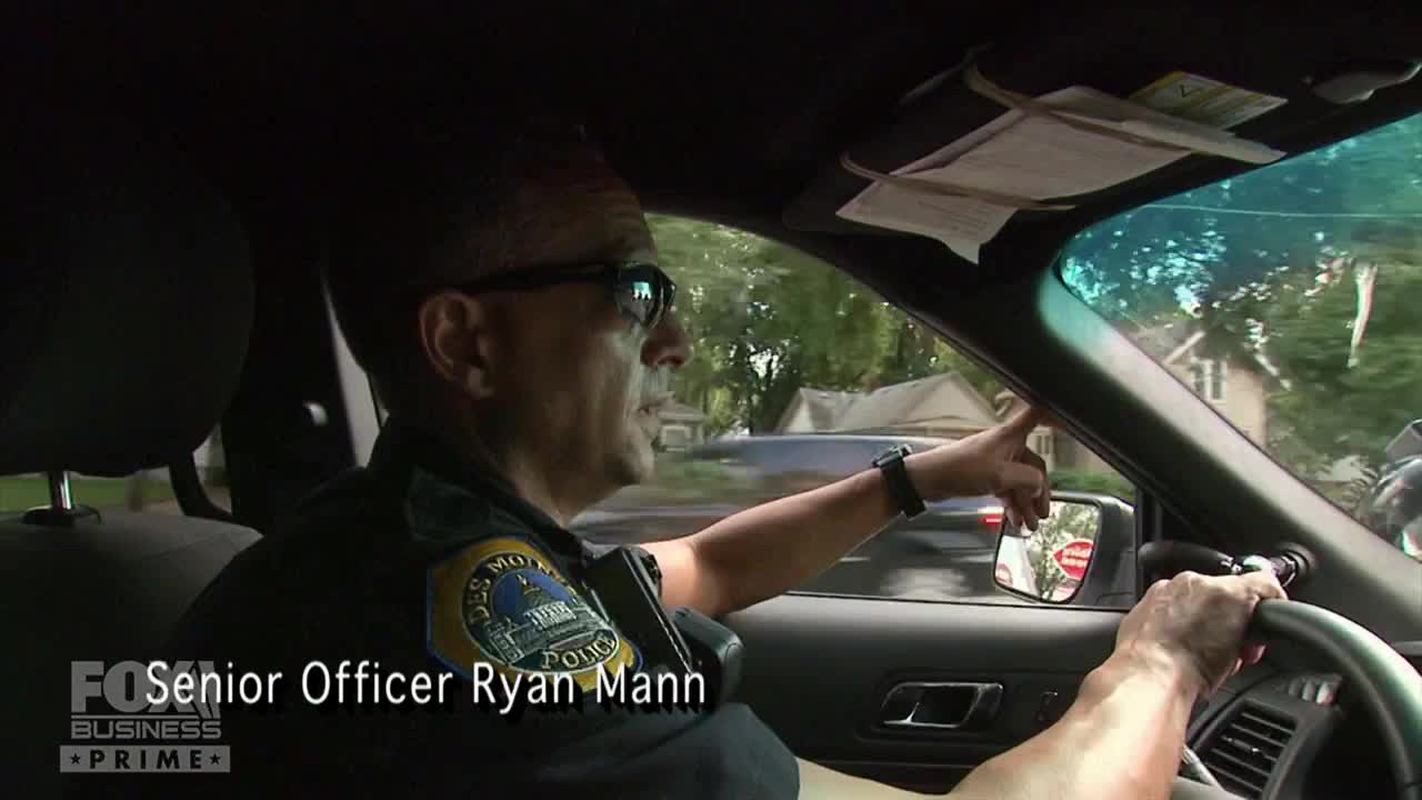 Senior Officer Ryan Mann, seen on Fox Business Network, points out the window while driving. The police officer, wearing sunglasses, is navigating a residential street in the United States.
