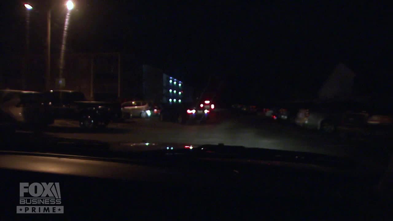 Streetlights illuminate a row of parked cars and a building with lit windows.  Headlights of vehicles are visible further down the road, likely heading toward the camera.
