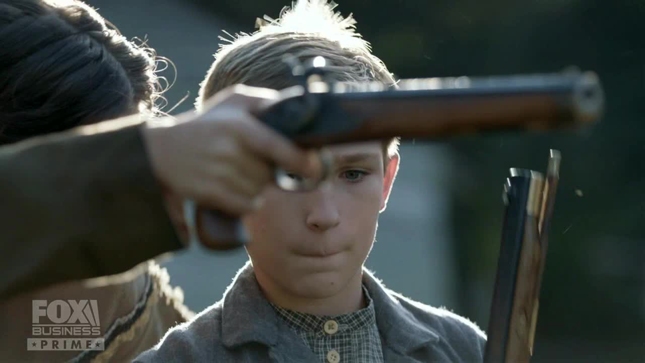 A young man stares forward as a rifle is aimed in his direction. The gun's barrel is pointed at the boy's face, and a second, broken rifle is held below.
