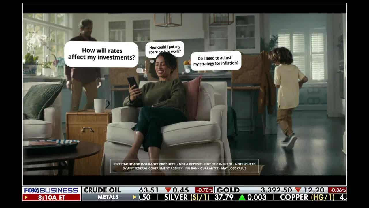 A woman sits in a living room, looking at her phone while thought bubbles float above her head. A child walks away from her, and a man stands in the background, the Fox Business Network ticker running at the bottom of the screen.
