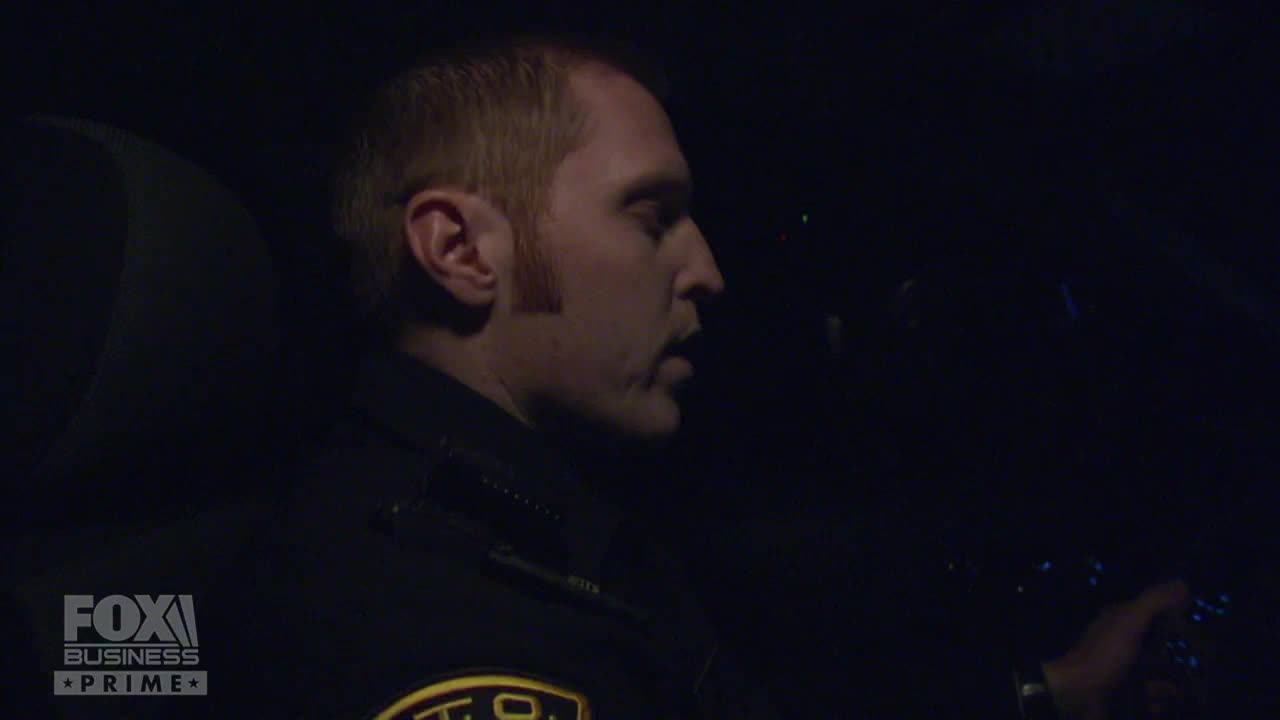 A man in a uniform, likely a police officer, is inside a vehicle, illuminated by the dashboard lights. He's looking down, with his hand on a control panel, as the Fox Business Network logo appears in the corner.
