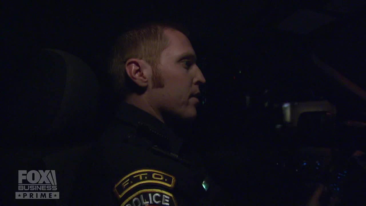 A police officer, illuminated by the interior lights of a car, speaks to someone off-camera. The Fox Business Network logo is visible in the lower left corner.
