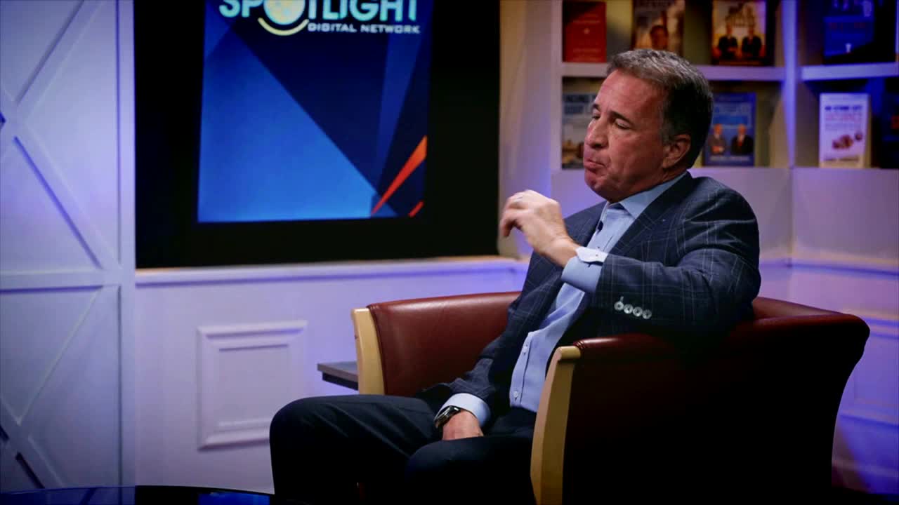 The man in the suit sits in a leather chair, thoughtfully touching his lower lip. Behind him, a screen displays the "Spotlight Digital Network" logo, likely on Fox Business Network.
