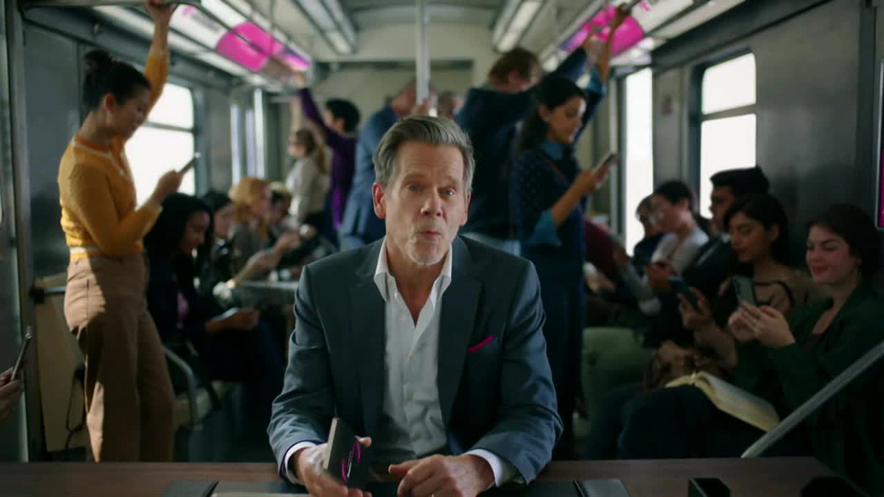 A man in a suit sits on a train, holding a phone, looking directly at the camera. Most of the other passengers are occupied with their phones, too.
