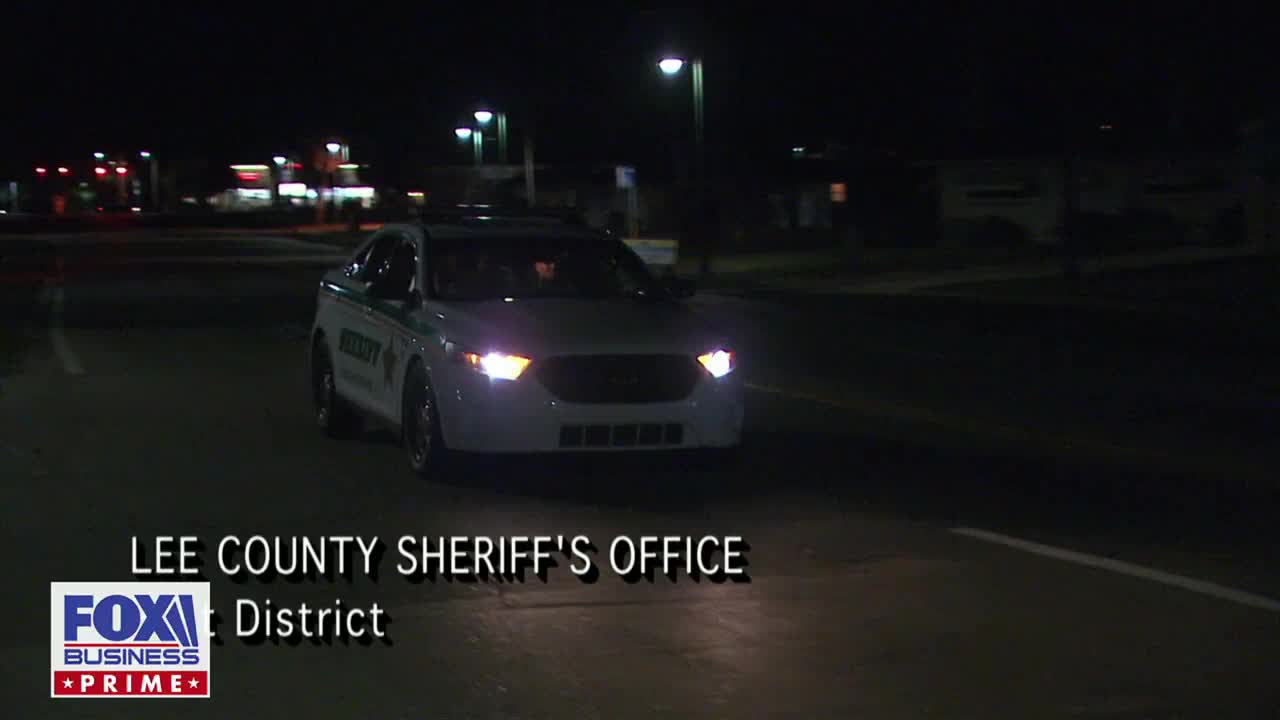 A Lee County Sheriff's Office patrol car, its headlights on, slowly approaches down a dark road. The scene is being broadcast on Fox Business Network.
