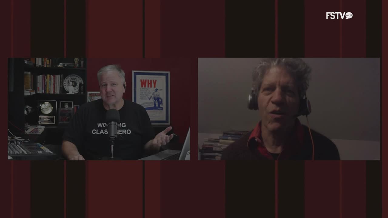 A man in a "Working Class Hero" t-shirt gestures while speaking into a microphone on Free Speech TV. Across the screen, another man wearing headphones is talking.
