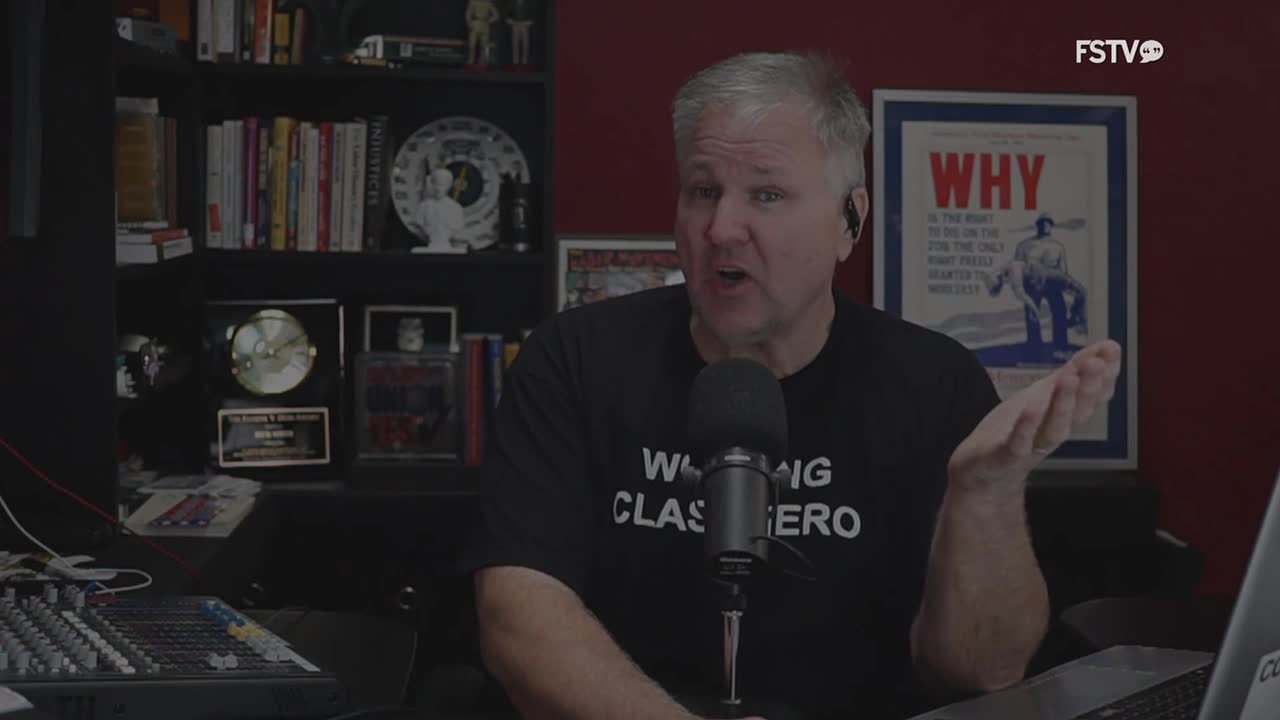 A man with graying hair, wearing a black t-shirt, gestures emphatically at the camera. He's speaking into a microphone, likely on Free Speech TV, with a bookshelf and framed poster behind him.
