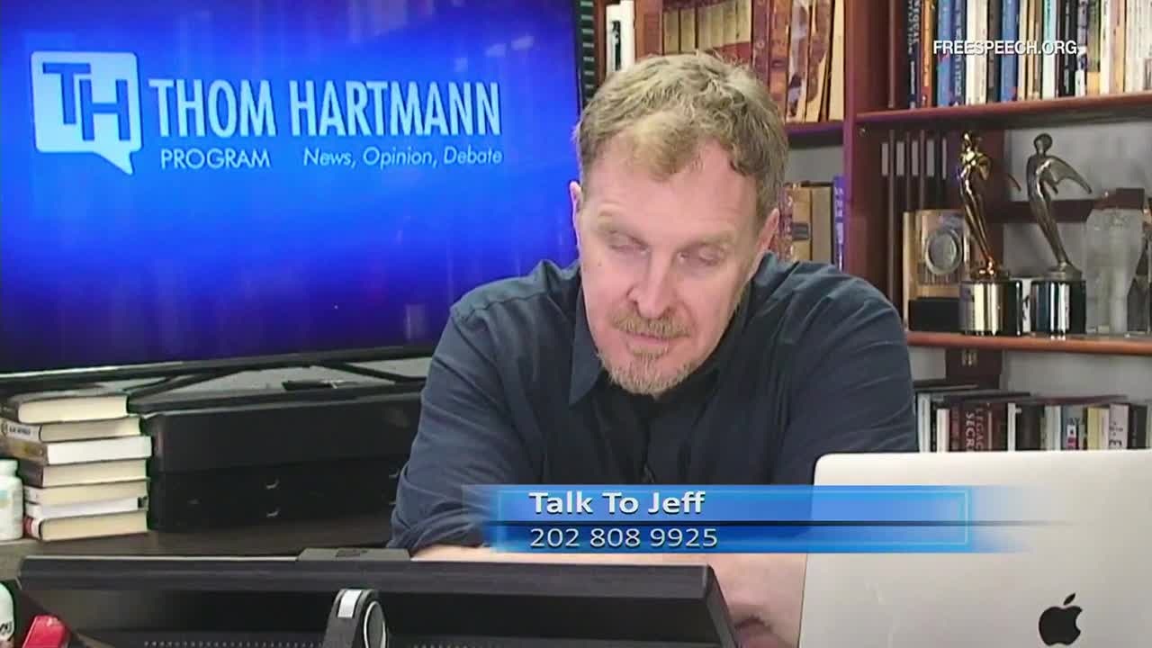 Thom Hartmann of Free Speech TV is looking down, likely at a computer, with a phone number displayed on the screen. Behind him, a bookshelf and a large monitor displaying his show's logo provide the backdrop.
