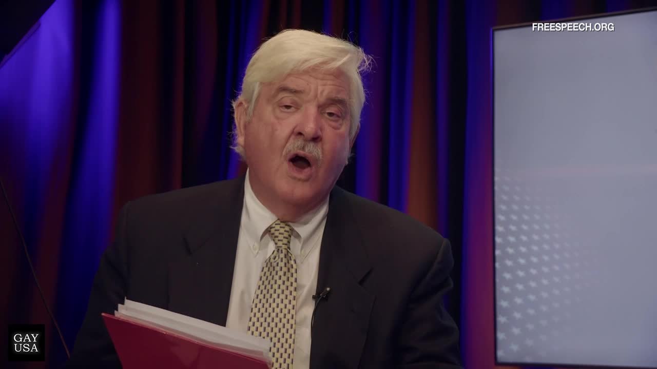 A man in a suit, tie, and a full mustache is speaking, his mouth open as if mid-sentence. He holds a red folder, and the Free Speech TV logo is visible on a screen behind him.
